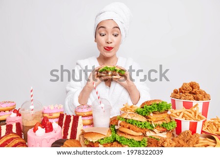 Hungry Asian woman licks red painted lips looks at tasty hamburger picks delicious snack break diet wears bathrobe and towel on head after taking shower chooses from yummy appetising fast food