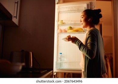 Hungry Asian Woman Taking Food Out Of The Fridge In The Kitchen At Night. Copy Space.