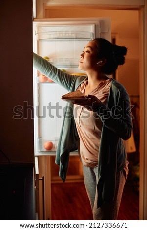 hungry Asian woman taking food from the refrigerator while having a snack in the evening at home. 