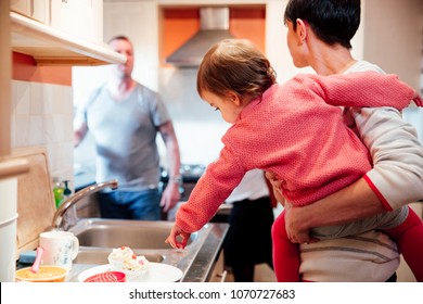 Hungry Baby Wants The Cupcakes That She Has Spotted On The Kitchen Worktop. She Is On Her Mother's Hip, Pointing To The Cakes But Her Parents Are Busy Talking To Eachother. 