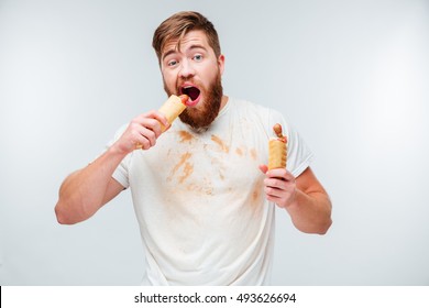 Hungry bearded man in dirty shirt biting two hotdogs isolated on white background