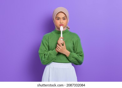 Hungry beautiful Asian woman in a green sweater holding a spoon in her mouth dream of a tasty meal isolated over purple background