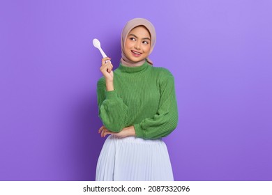 Hungry Beautiful Asian Woman In Green Sweater Holding Spoon Think Of Tasty Food Isolated Over Purple Background