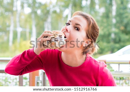 Hungry beautiful young woman is biting, eating juicy hamburger outdoor at summer day, looking at camera. Girl eating fast food, burger with appetite. Street junk unhealthy food concept. Gain mass, fat