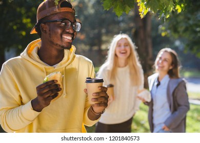 Hungry Black Afro Hipster Man Student Having Lunch, Hold Sandwich And Coffee To Go While Standing Outdoor With Two Female On Blurred Background In Sunny Autumn Day. Get-together Meeting Concept.