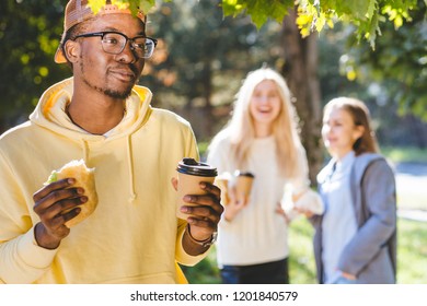 Hungry Black Afro Hipster Man Student Having Lunch, Hold Sandwich And Coffee To Go While Standing Outdoor With Two Female On Blurred Background In Sunny Autumn Day. Get-together Meeting Concept.