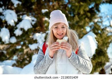 Hungry Blonde Woman Eating Sandwich During Hiking Break In Snowy Mountain. Mature Female Enjoying In Snack Outdoors In Cold Sunny Winter Day, Near Fir Tree. Weekend Active Lifestyle. Copy Space