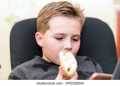 A Hungry Boy Eating A Hot Dog At Home Sit While Playing With The Smartphone. Indoors Shot.Closeup.