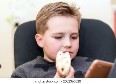 A Hungry Boy Eating A Hot Dog At Home Sit While Playing With The Smartphone. Indoors Shot.Closeup.