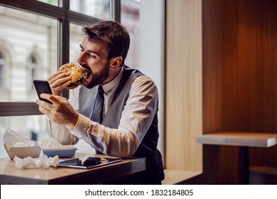 Hungry Businessman Sitting On Lunch Break In Fast Food Restaurant, Eating Cheese Burger And Using Smart Phone.