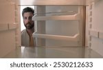 Hungry Caucasian bachelor man looking for food in empty fridge finding no meal products feeling sad disappointed poor unemployment need delivery service POV point of view from inside refrigerator