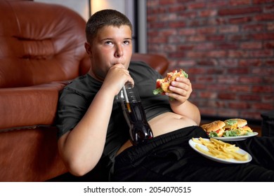 Hungry Caucasian Fat Boy Is Eating Junk Food At Home While Watching Tv. Unhealthy Fast Food. Overweight Teenage Child Sit On Floor Looking At Side, Eating Unhealthy Food. Lifestyle, Overeating