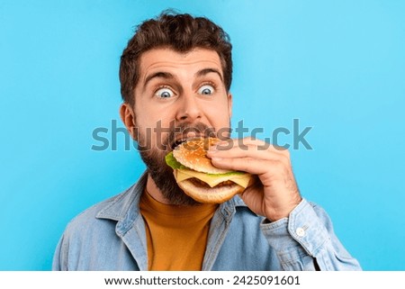 Hungry caucasian man on blue background excitedly biting into burger, revealing pleasure of indulging in junk food. Headshot of excited guy eating cheeseburger. Fast food cravings concept