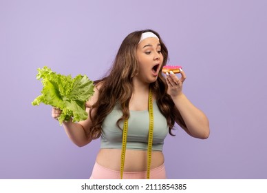 Hungry Caucasian Plus Size Young Female In Sportswear Eat Donut And Hold Green Salad Isolated On Purple Background. Junk Food And Excess Weight, Diet And Exercise, Sweet Addiction And Proper Nutrition