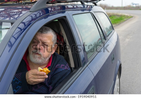 Hungry\
Caucasian senior driver eating patty with apple inside his car on\
short stop on roadside while traveling in\
Ukraine