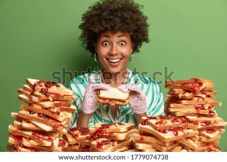 Hungry cheerful woman eats delicious snack, looks positively at camera, has good appetite and gluttony, wears elegant dress and gloves, poses near many bread toasts, isolated on green studio wall