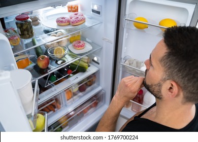 Hungry Confused Man Looking In Open Fridge In Kitchen