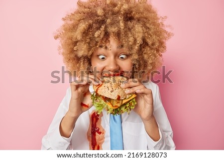 Hungry curly haired woman eats delicious sandwich enjoys fast food has unhealthy nutrition smeared with ketchup dressed in formal clothes isolated over pink background. Binge eating concept.