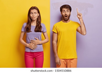 Hungry dark haired woman touches stomach, wants to eat something tasty, wears purple t shirt and pink trousers, impressed unshaven man in casual wear points index finger above, shows direction up