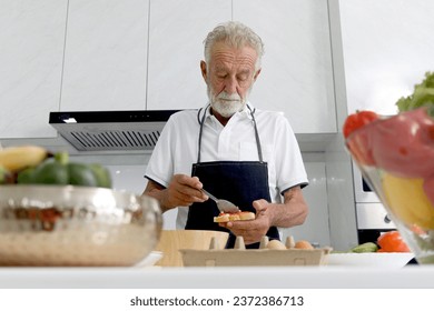 Hungry elderly man wearing apron at kitchen counter with colorful fresh vegetables and fruits. Senior man holding bread for making sandwich, mature grandfather enjoys cooking meal by himself at home. - Powered by Shutterstock