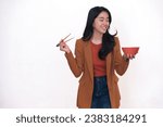 Hungry facial expression of a young Asian woman sniffing her food in a red ceramic bowl, while other hand holding a pair of chopsticks