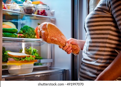 Hungry Fat Woman Holding A Big Turkey Leg In His Hands And Standing Next To The Open Fridge. Unhealthy Food.