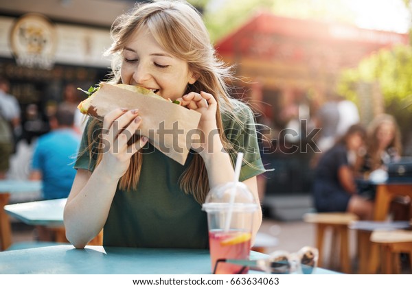 Hungry freckled blonde girl eating taco on a\
food court on a sunny summer day in\
park.