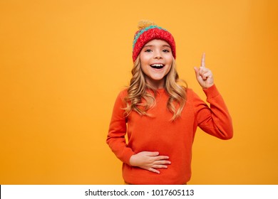 Hungry Happy Young Girl In Sweater And Hat Holding Her Tummy And Having Idea While Looking At The Camera Over Orange Background