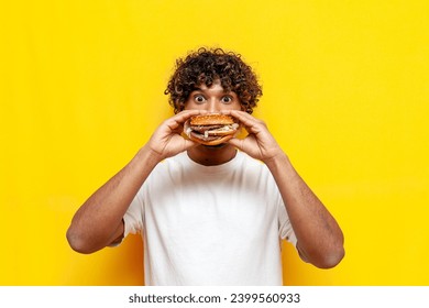 hungry indian guy eats a delicious burger on a yellow isolated background and is surprised, young indian man bites fast food and eats unhealthy food