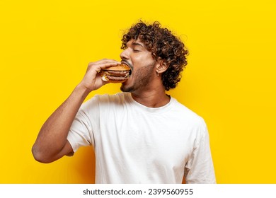 hungry indian guy eats a delicious burger on a yellow isolated background and is surprised, young indian man bites fast food and eats unhealthy food - Powered by Shutterstock