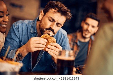 Hombre hambriento comiendo hamburguesa mientras se reúne con amigos en un pub.