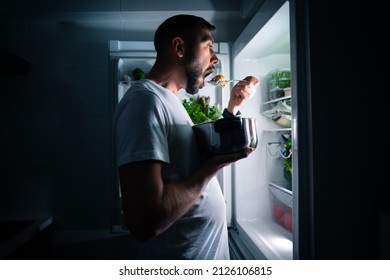 Hungry man eating food at night from open fridge. Man taking midnight snack from refrigerator