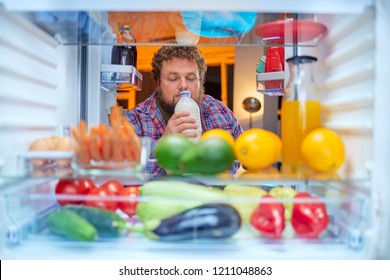 Hungry Man Standing In Front Of Fridge And Smelling Milk Late At Night. Eating Disorder, Unhealthy Eating. Picture Taken From The Inside Of Fridge.