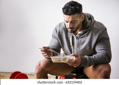 Hungry muscular young man gulping down food looking at plastic container without pausing as he takes another mouthful