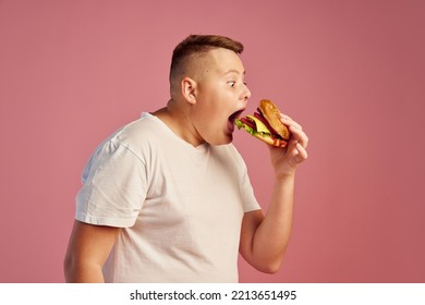 Hungry Overweight Teen Boy In White T-shirt Biting Delicious Hamburger Isolated On Pink Background. Fast Food, Taste, Body Positive, Emotions And Facial Expressions. Copy Space For Ad