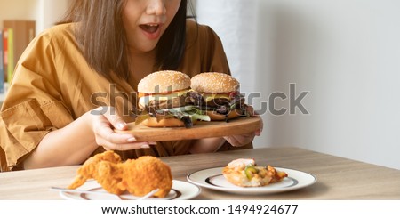 Hungry overweight woman holding hamburger on wooden plate, Fried chicken and Pizza on table .Concept of binge eating disorder (BED). Stock fotó © 