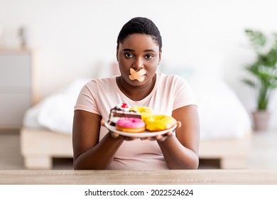 Hungry Plus Size Black Woman Having Strict Weight Loss Diet, Wearing Adhesive Bandages On Her Mouth, Holding Plate With Sweets, Refusing To Eat Desserts. Slimming Restrictions Concept