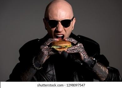 Hungry Tattooed Man In Leather Jacket And Sunglasses Eating Hamburger Isolated On Grey