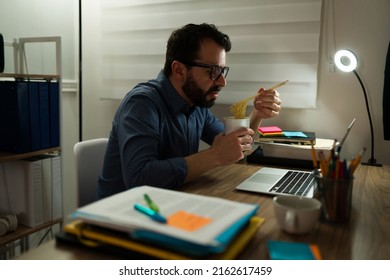 Hungry Tired Man Working Very Late At Night In The Office And Eating Noodles At His Desk 