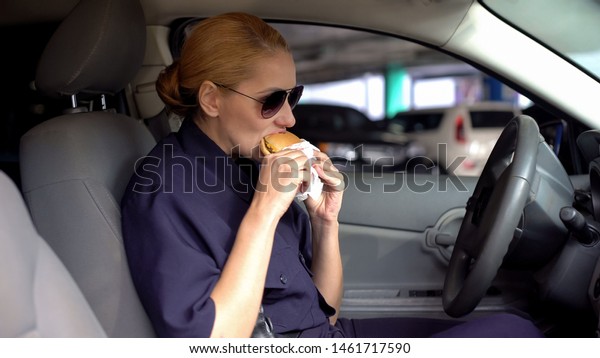 Hungry woman cop eating burger sitting in police car\
in parking lot, junk\
food