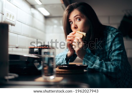 Hungry Woman Eating a Sandwich at Night in the Kitchen. Young person stress eating during nighttime having a late snack 
