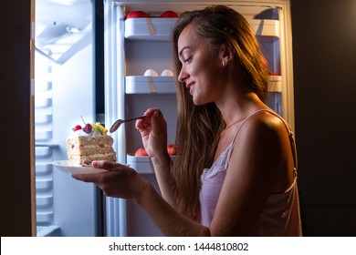 Hungry woman in pajamas eating sweet cake at night near fridge. Stop diet and gain extra pounds due to high carbs junk food and unhealthy night eating
