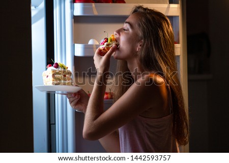 Hungry woman in pajamas eating sweet cakes at night near refrigerator. Stop diet and gain extra pounds due to high carbs food and unhealthy night eating