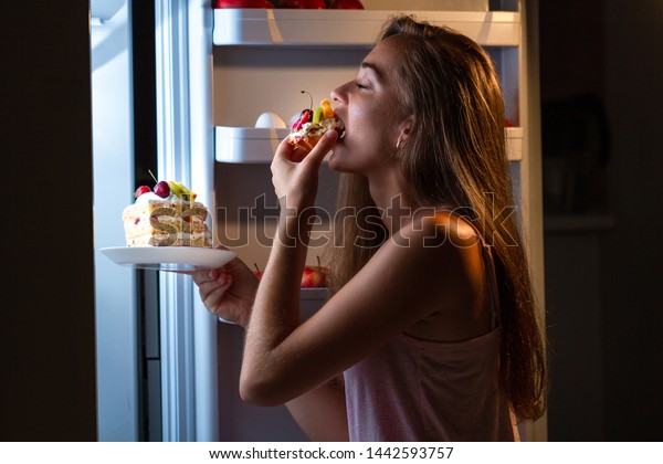 Hungry woman in pajamas eating\
sweet cakes at night near refrigerator. Stop diet and gain extra\
pounds due to high carbs food and unhealthy night\
eating