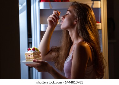 Hungry woman in pajamas enjoys sweet cake at night near refrigerator. Stop diet and gain extra pounds due to carbs food and unhealthy eating 