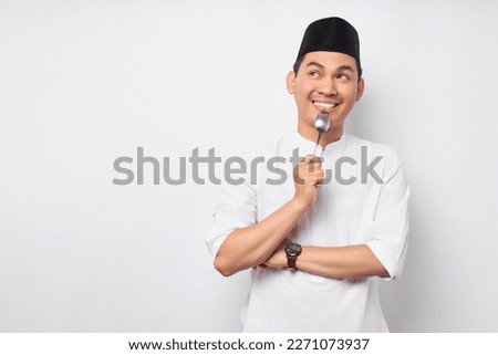 Hungry young Asian Muslim man in Arabic clothes with spoon in her mouth and looking aside, dreamy delicious food to break fast isolated on white background. People religious Islamic lifestyle concept