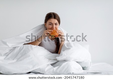 Hungry young female greedily eating burger on bed. Eating disorder, consolation with overeating and gluttony, obesity after mental depression, loss substitution with unhealthy food consumption problem