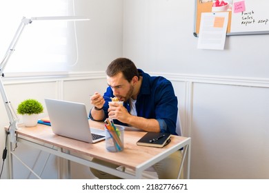 Hungry Young Man Eating Lunch At His Desk While Looking At The Computer And Doing Office Work  