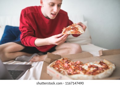 Hungry young man in a jersey sweatshirt sits at home on a bed with a pizza box and a laptop, holds a piece of pizza in his hands and is about to eat it. Teen eats at home on a pizza bed with delivery - Powered by Shutterstock