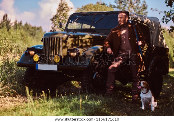Hunter\
in elegant clothes holds a shotgun and standing together with his\
beagle dog near a retro military car in a\
forest.